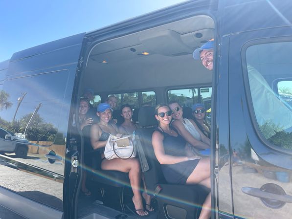 Smiling group of young adults seated in a black passenger van with the sliding door open on a sunny beachside street, palm trees and blue sky reflected on the van.