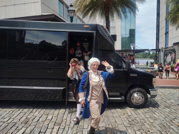 Person in blue colonial costume waving while stepping from a black shuttle van with companions in period dress on a cobblestone waterfront plaza lined with palm trees and modern buildings.