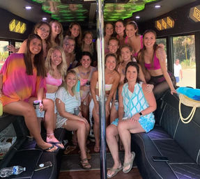 Smiling group of women in colorful swimsuits and summer outfits posing inside a party bus around a center pole, holding drinks, palm trees visible outside - beachside celebration.