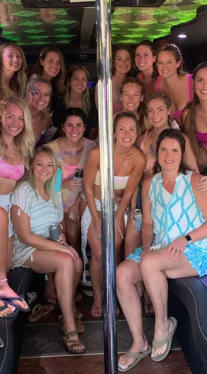 Smiling group of women in colorful swimsuits and summer outfits posing inside a party bus around a center pole, holding drinks, palm trees visible outside - beachside celebration.