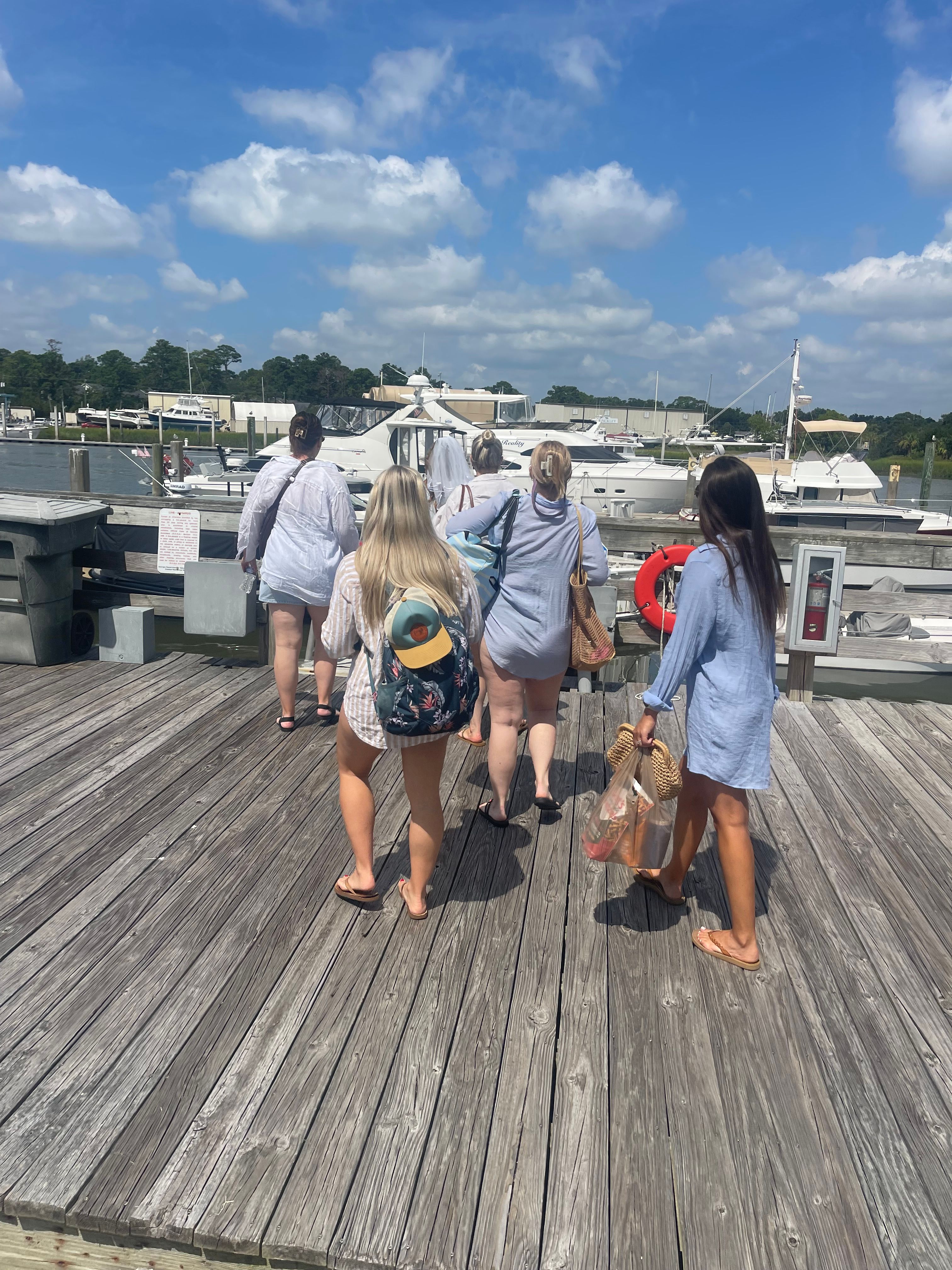 Five friends in light summer cover-ups and flip-flops walking on a weathered wooden marina dock toward moored yachts and boats under a sunny blue sky