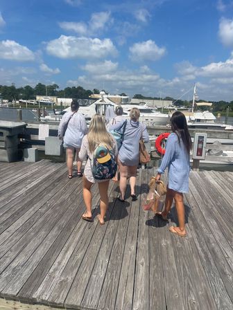 Five friends in light summer cover-ups and flip-flops walking on a weathered wooden marina dock toward moored yachts and boats under a sunny blue sky