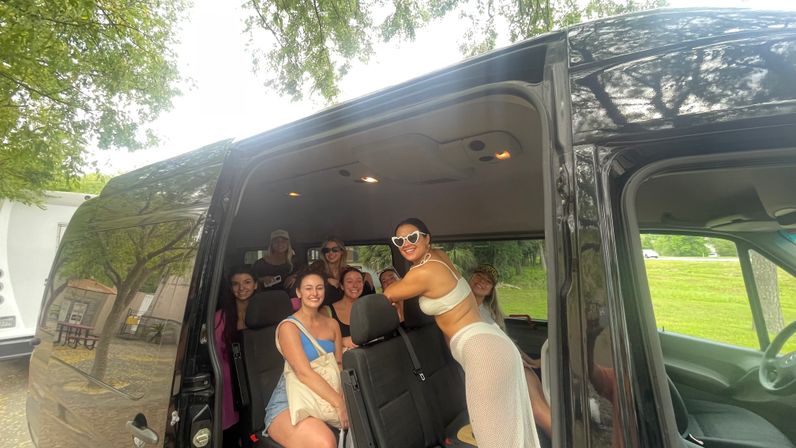 Group of smiling women inside a black passenger van with the sliding door open, parked under trees beside a grassy park; one woman in a white mesh outfit and heart-shaped sunglasses leans on a seat while others sit and pose for a fun road-trip photo.