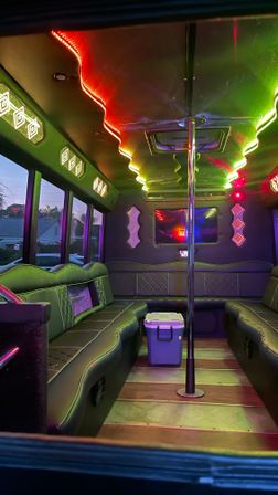 Party bus interior with leather bench seating, center dance pole, multicolor LED ceiling lights and a cooler, suburban evening view through side windows.