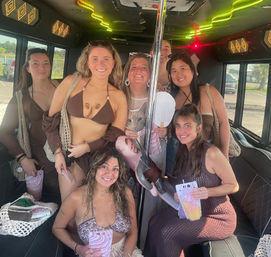 Seven women in swimsuits smiling inside a neon-lit party bus around a chrome pole, holding drinks and striped gift bags while parked in a sunny lot
