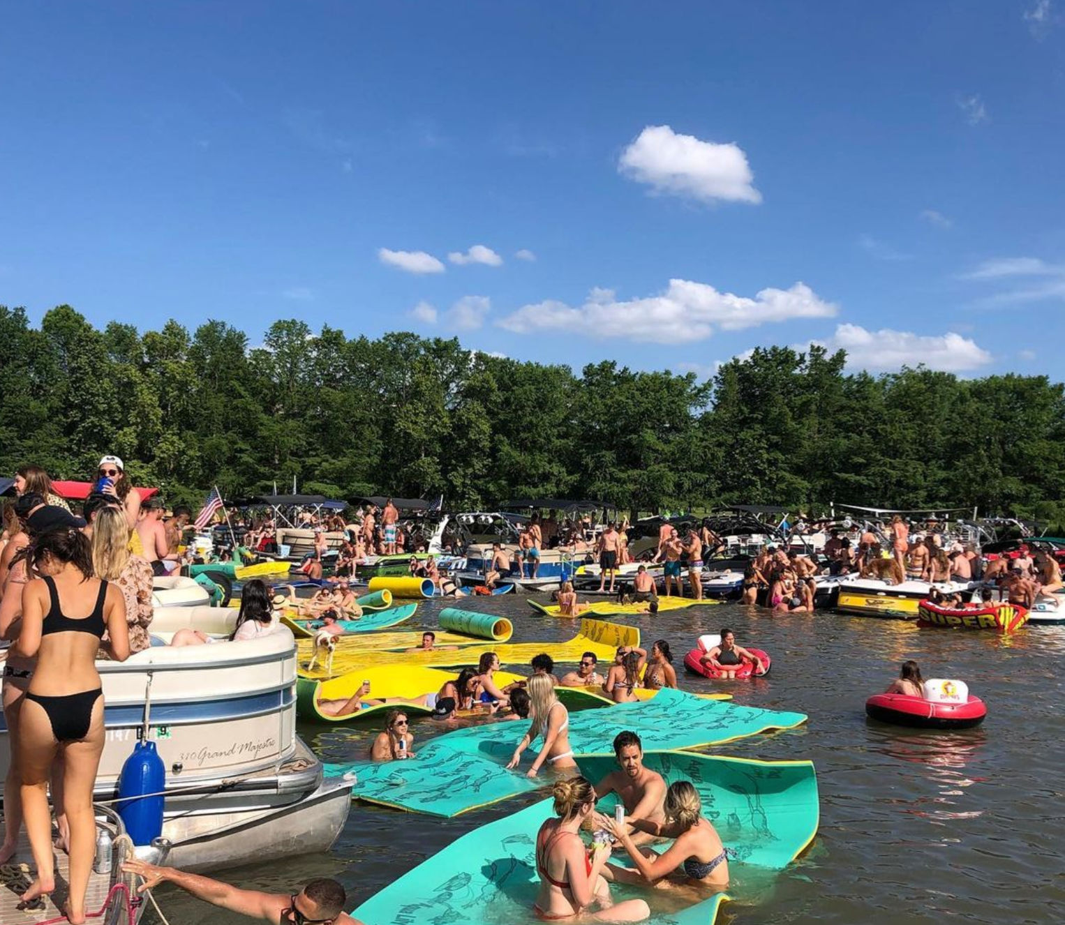 Crowded summer lake party with boats, inflatable tubes and bright foam float mats on the water under a sunny blue sky and tree-lined shore.