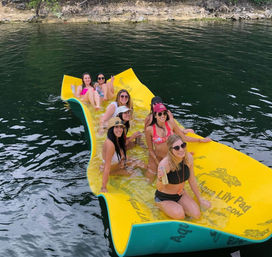 Group of women in swimsuits enjoying summer on a bright yellow floating pad in a lake near a wooded shoreline, smiling and holding drinks.