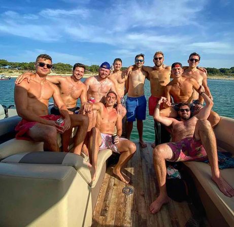 Group of shirtless friends on a pontoon boat enjoying a summer lake day with sunglasses and drinks, wooden deck, shoreline and blue sky in the background