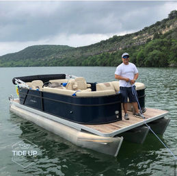 Person in a white T-shirt, cap and sunglasses tying a rope on the bow of a navy pontoon boat with beige seats on a calm lake, green tree-lined cliffs and cloudy sky in the background.