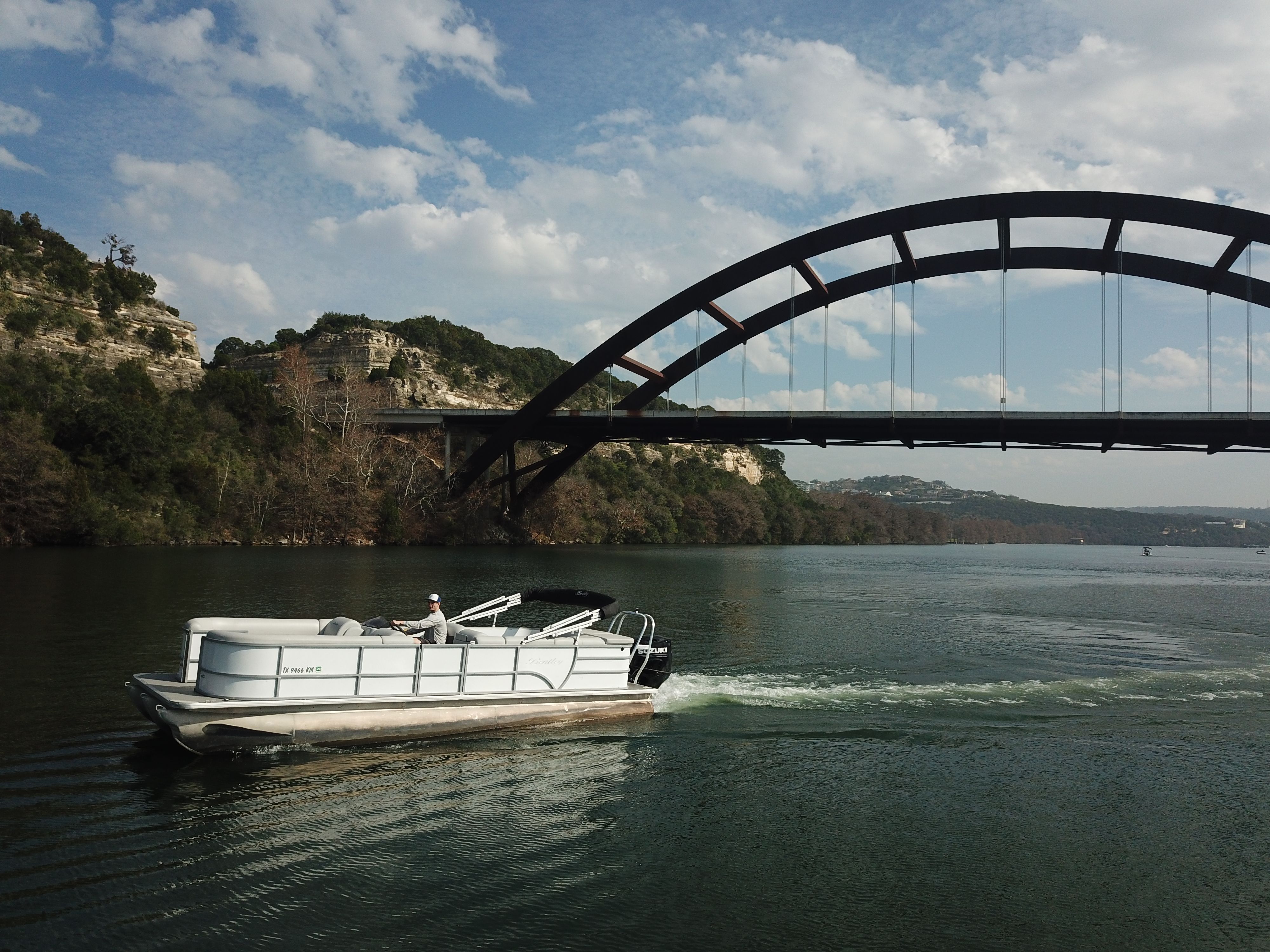 Pontoon boat cruising on a calm lake beneath a large arched steel bridge with limestone cliffs and a partly cloudy sky