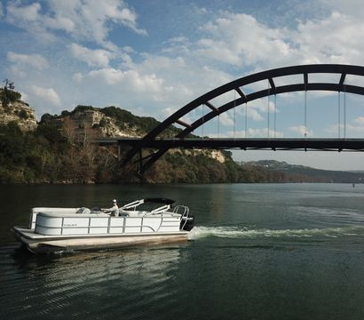 Pontoon boat cruising on a calm lake beneath a large arched steel bridge with limestone cliffs and a partly cloudy sky