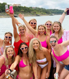 Cheerful bachelorette party on a lake boat — group of women in pink swimsuits and heart-shaped sunglasses, bride-to-be in white 'bride' swimsuit, smiling and raising drinks with wooded shoreline behind them