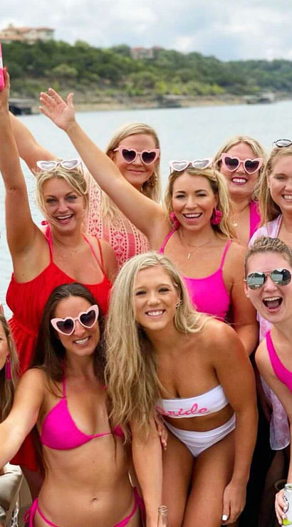 Cheerful bachelorette party on a lake boat — group of women in pink swimsuits and heart-shaped sunglasses, bride-to-be in white 'bride' swimsuit, smiling and raising drinks with wooded shoreline behind them