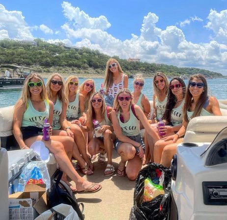 Group of friends on a pontoon boat enjoying a sunny lake day, wearing matching tank tops and colorful heart-shaped sunglasses while holding drinks, with tree-lined shoreline and puffy clouds overhead.