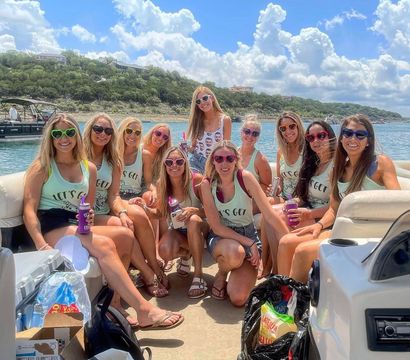 Group of friends on a pontoon boat enjoying a sunny lake day, wearing matching tank tops and colorful heart-shaped sunglasses while holding drinks, with tree-lined shoreline and puffy clouds overhead.