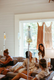 IV drip bag in focus in a sunlit living room as a group of women relax on sofas in robes with drinks, dresses hanging in the window — at-home wellness and pre-event pampering scene
