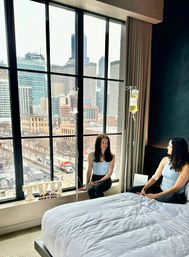 Two women in workout tops receiving colorful IV vitamin drips while relaxing in a hotel room by a floor-to-ceiling window with a downtown high-rise skyline view.