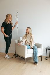 Smiling healthcare provider holding an IV pole while a relaxed woman receives IV therapy in a bright, cozy wellness clinic lounge.