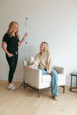 Smiling healthcare provider holding an IV pole while a relaxed woman receives IV therapy in a bright, cozy wellness clinic lounge.