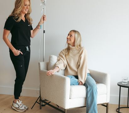 Smiling healthcare provider holding an IV pole while a relaxed woman receives IV therapy in a bright, cozy wellness clinic lounge.