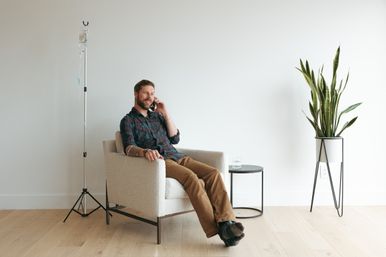 Relaxed man on phone receiving IV drip in a bright minimalist room with a small side table, glass of water, and tall potted snake plant.
