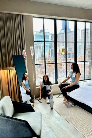Two women in athleisure receiving IV hydration drips while relaxing in a modern downtown hotel room with floor-to-ceiling windows and an urban skyline view