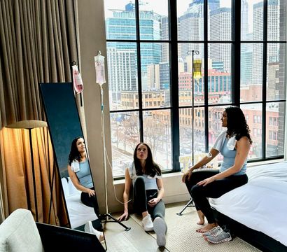Two women in athleisure receiving IV hydration drips while relaxing in a modern downtown hotel room with floor-to-ceiling windows and an urban skyline view
