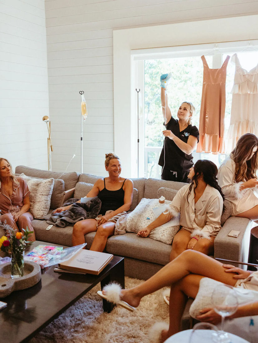 Women relaxing in a bright living room at an at-home spa party — wearing pajamas and fluffy slippers, sipping drinks and receiving IV hydration drips while dresses hang by the window.