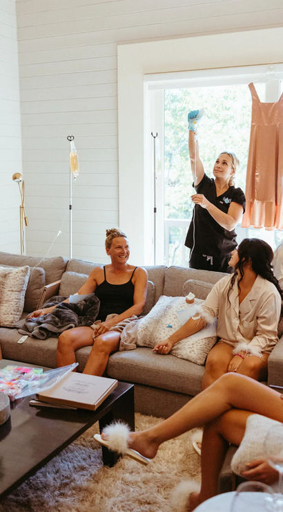 Women relaxing in a bright living room at an at-home spa party — wearing pajamas and fluffy slippers, sipping drinks and receiving IV hydration drips while dresses hang by the window.