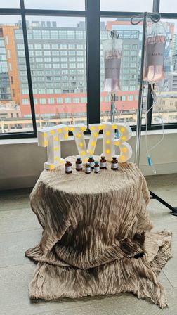 IV vitamin therapy display in a downtown loft — LED marquee letters, small vitamin vials on a draped beige table and two pink vitamin IV bags hanging by a window with city buildings outside.