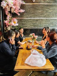 Friends chatting at a wooden communal table in a cozy restaurant with pink cherry blossom decor, stone accent wall and a small vase of roses