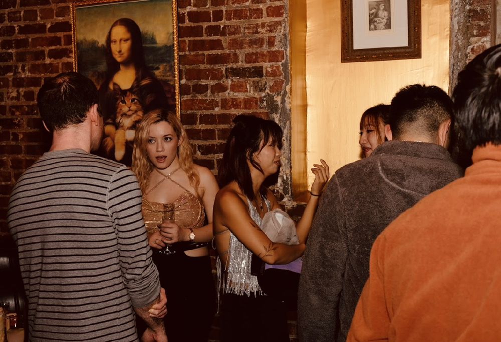 Young adults chatting and holding drinks in a dim, brick-walled downtown bar with a playful Mona Lisa-style portrait featuring a cat on the wall — lively nightlife scene.