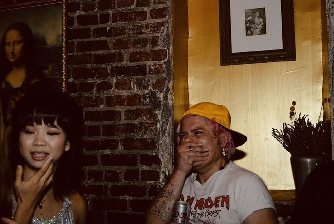 Two friends laughing in a dim downtown bar with exposed brick and a framed Mona Lisa print; one in a yellow cap covers their mouth holding a can while the other in a sparkly top touches her chin, dried flowers in a vase nearby.