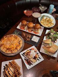 Restaurant table spread in a cozy booth with comfort food and cocktails — cheesy pizza, three mini sliders, seasoned fries, grilled skewers with dip, panini and salad, dessert waffles and chocolate-drizzled crepes, plus colorful craft cocktails.