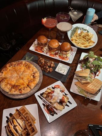 Restaurant table spread in a cozy booth with comfort food and cocktails — cheesy pizza, three mini sliders, seasoned fries, grilled skewers with dip, panini and salad, dessert waffles and chocolate-drizzled crepes, plus colorful craft cocktails.