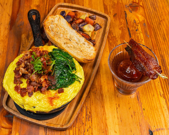Brunch skillet omelette topped with crispy bacon and sautéed spinach, served with toasted bread and roasted potatoes on a wooden tray beside a cocktail garnished with candied bacon on a rustic wooden table.
