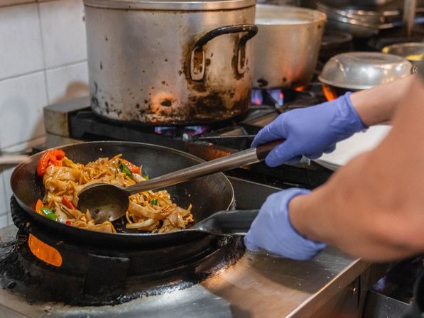 Gloved hands stir-frying wide noodles with peppers and greens in a wok over a gas stove in a busy commercial kitchen.