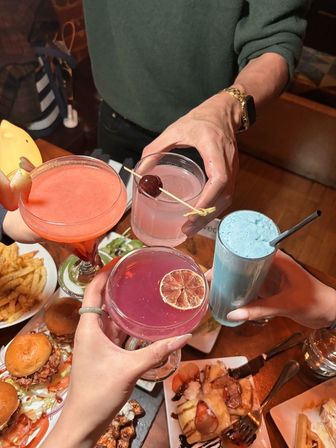 Cheers at a restaurant table: four hands clinking colorful cocktails and a blue milkshake over a wooden table with sliders, fries and dessert, pink and red drinks garnished with citrus and a cherry.