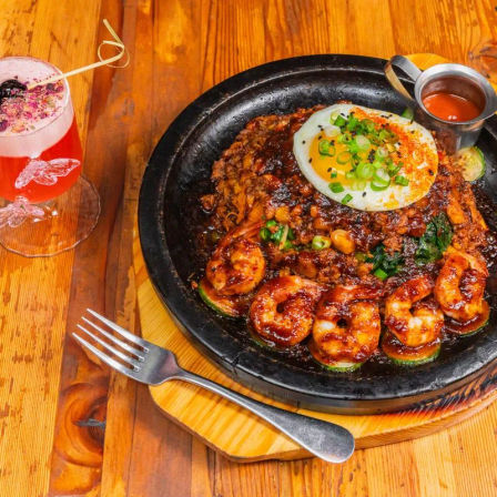 Sizzling cast-iron skillet of spicy glazed shrimp and seasoned minced meat topped with a sunny-side egg and chopped scallions, served with dipping sauce and a pink floral cocktail on a wooden table.