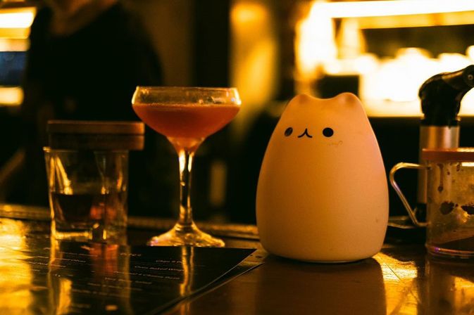 Cute cat-shaped ceramic lamp beside a pink cocktail in a coupe glass on a warm, dimly lit bar counter with glass jars and cozy cocktail bar ambiance