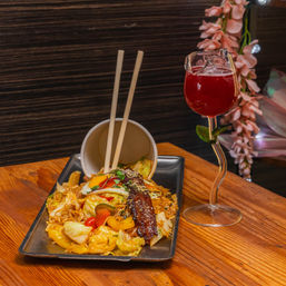 Asian-fusion stir-fried noodles with cabbage, bell peppers, a fried egg and glazed pork belly on a rectangular plate with chopsticks, paired with a red cocktail in a stemmed glass on a wooden table in a cozy restaurant setting