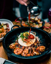 Close-up of a sizzling stone bowl of rice topped with caramelized shrimp, a sunny-side-up egg, chopped scallions and spicy red sauce being poured over it.