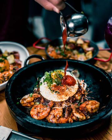 Close-up of a sizzling stone bowl of rice topped with caramelized shrimp, a sunny-side-up egg, chopped scallions and spicy red sauce being poured over it.