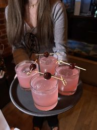 Four rosy pink cocktails in short rocks glasses, each garnished with a cherry on a bamboo skewer, served on a black tray in a cozy bar setting.