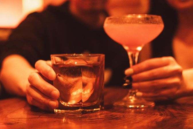 Two hands holding cocktails at a wooden bar—a whiskey on the rocks in a lowball glass and a pink coupe cocktail—bathed in warm amber bar lighting.