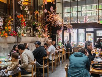 Light-filled urban cafe with cascading pink and orange floral decor, communal wooden tables and patrons enjoying brunch by large street-facing windows.