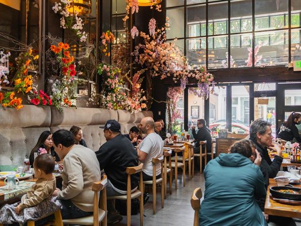 Light-filled urban cafe with cascading pink and orange floral decor, communal wooden tables and patrons enjoying brunch by large street-facing windows.