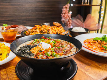 Black bowl of savory rice porridge topped with seaweed, crunchy bits, scallions and a soft-poached egg, surrounded by colorful Asian-inspired dishes on a wooden restaurant table — cozy comfort-food spread.