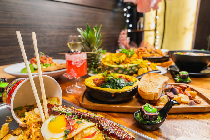 Asian-fusion feast on a wooden restaurant table: chopsticks in a noodle cup with sunny-side egg and glazed eel, sushi bite on a spoon, baked spinach skillet, toast and roasted potatoes, pineapple boat stir-fry, pink cocktail and wine glass