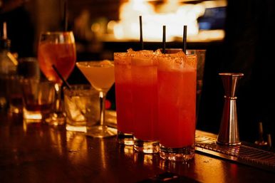 Three tall red-orange cocktails with crushed ice and straws on a dim wooden bar counter, assorted mixed drinks and a jigger in the blurred background — cozy urban nightlife cocktail scene
